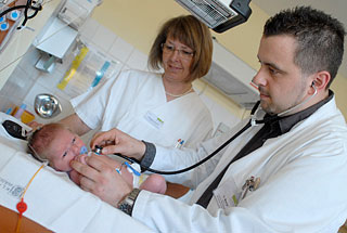 Oberarzt Stangl untersucht ein Neugeborenes mit einem Steoskop, eine Schwester schaut zu